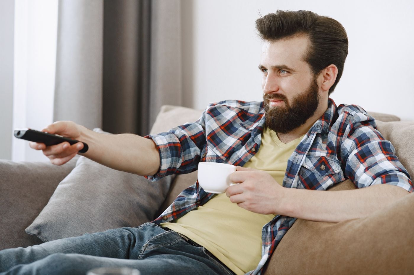 Caucasian man with a beard relaxing at home, holding a cup and watching TV on a comfortable sofa.