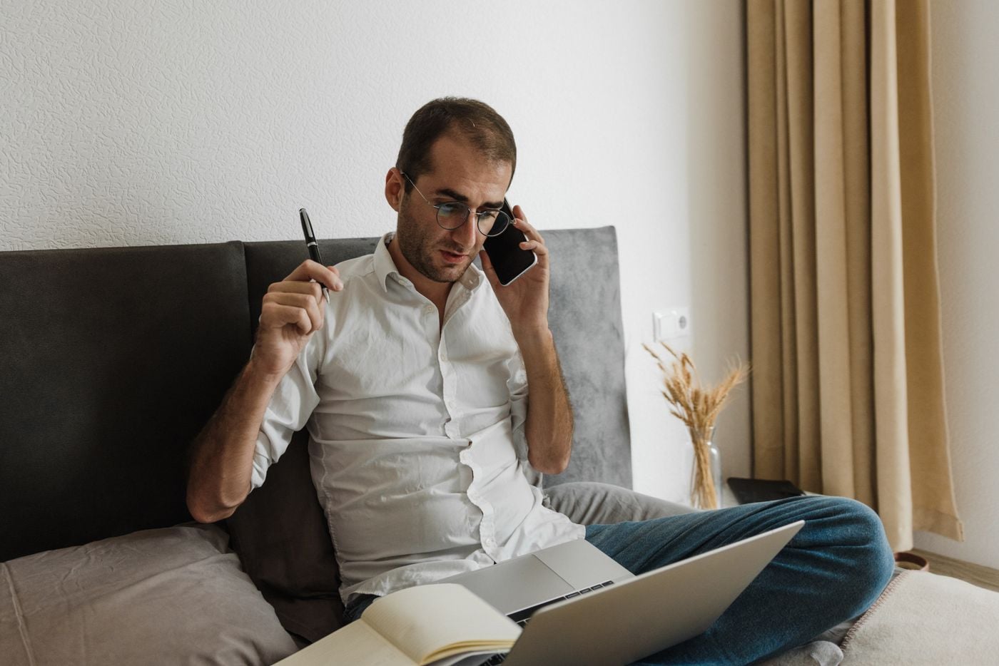 Adult male multitasking at home office, using laptop and smartphone while sitting on bed.